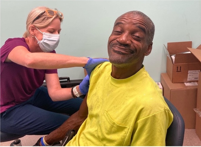 The image titled "Patient getting vaccinated at VAXXED & BOOSTED event" shows a man receiving a COVID-19 vaccine during a community outreach campaign aimed at increasing vaccination rates in West Greenville, North Carolina. The patient, smiling while getting vaccinated, reflects the success of the event in creating a welcoming and positive atmosphere. The initiative, as described in the article, used local ambassadors and accessible locations to combat vaccine hesitancy and educate the community about the benefits of vaccination​.