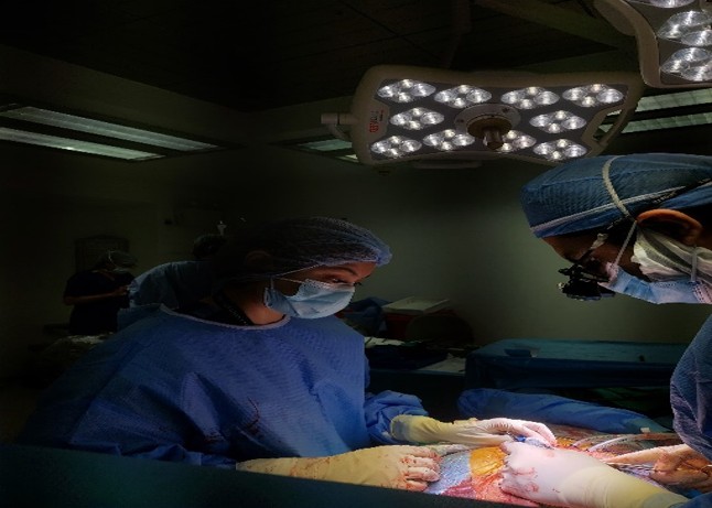 The photograph depicts a surgical team performing an operation under sterile conditions in a well-equipped operating room. Two surgeons, fully dressed in surgical gowns, masks, and gloves, are focused on the open surgical field, illuminated by an overhead surgical light. The environment reflects standard aseptic technique, with clear teamwork and attention to detail during a complex procedure.
