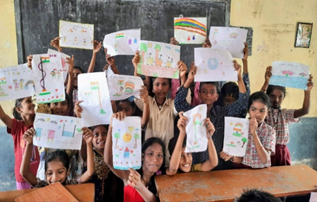 The image shows a group of smiling schoolchildren proudly displaying their colorful drawings inside a classroom. The children are gathered in front of a blackboard, enthusiastically holding up artwork that features houses, trees, rainbows, and national flags. The vibrant atmosphere reflects joy, creativity, and engagement in a school activity, highlighting the positive impact of educational and artistic expression in young learners.